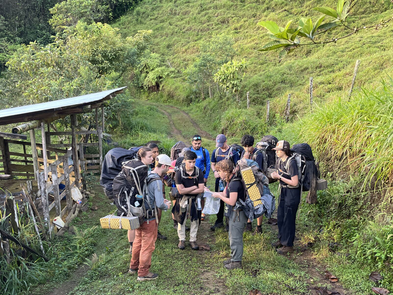 A group of people are gathered on a grassy path, some with large backpacks, possibly preparing for a hike or trek. They appear to be consulting a map or instructions. The setting is a rural, possibly mountainous area with lush green vegetation and a dirt path leading uphill. A wooden structure with a roof is visible to the left.
